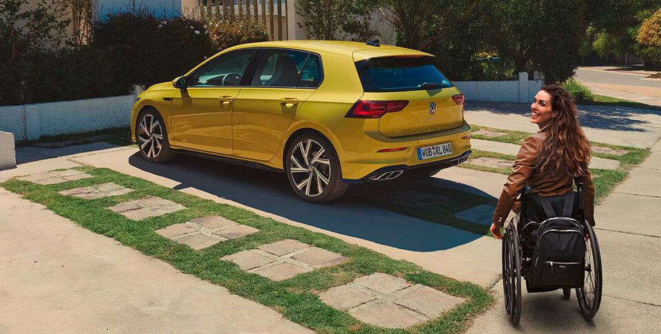 Eine Frau in Rollstuhl nähert sich einem parkenden gelben VW Golf.